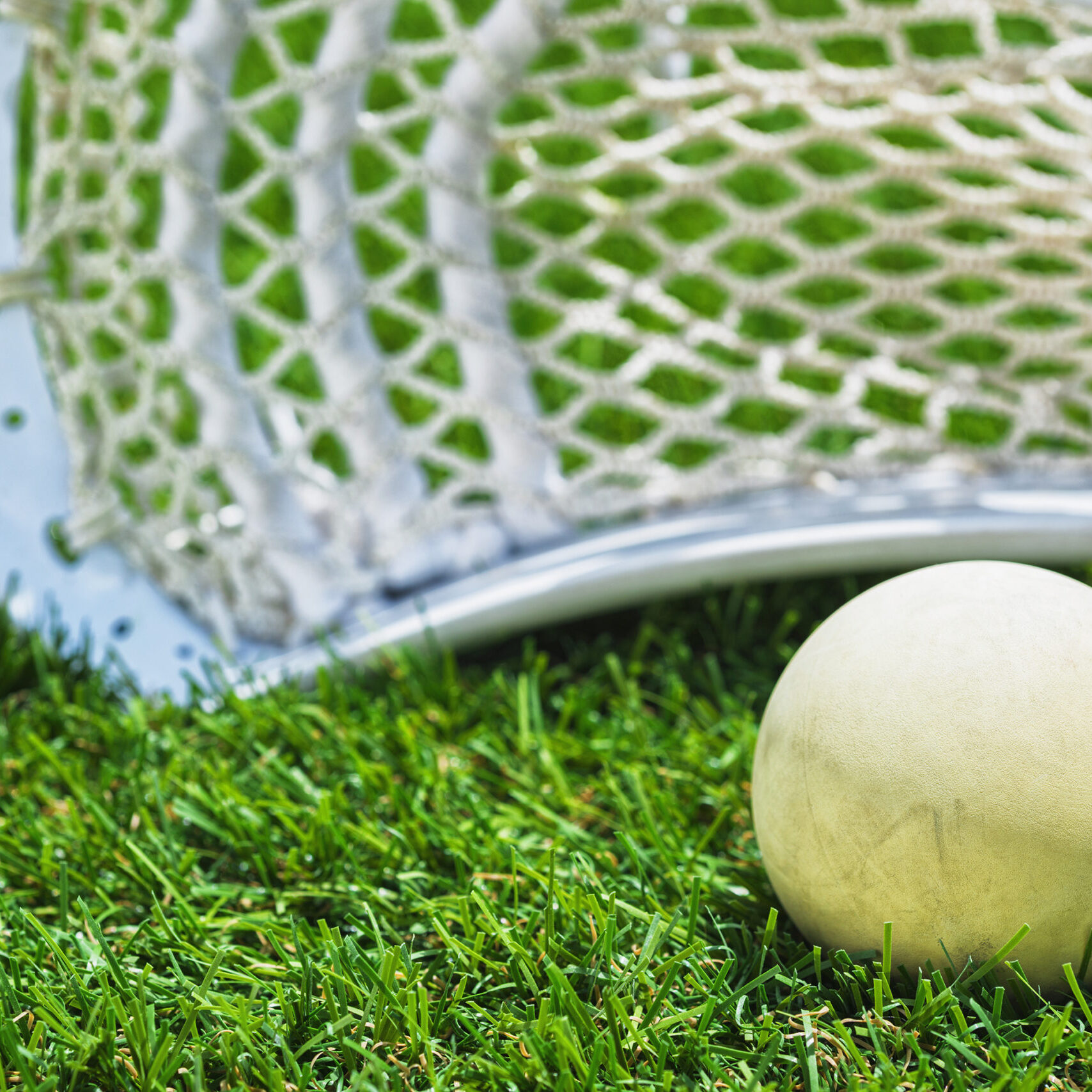 Low angle view of a white Lacross ball sitting in front of a Lacrosse stick with white shooting string on artificial turf in the mid day sun